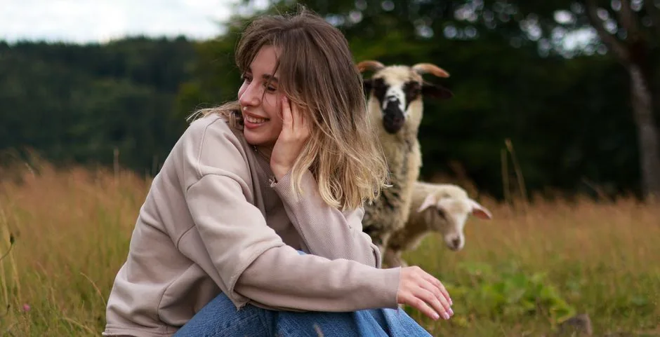 A woman smiles while sitting in a meadow with sheep in the background.