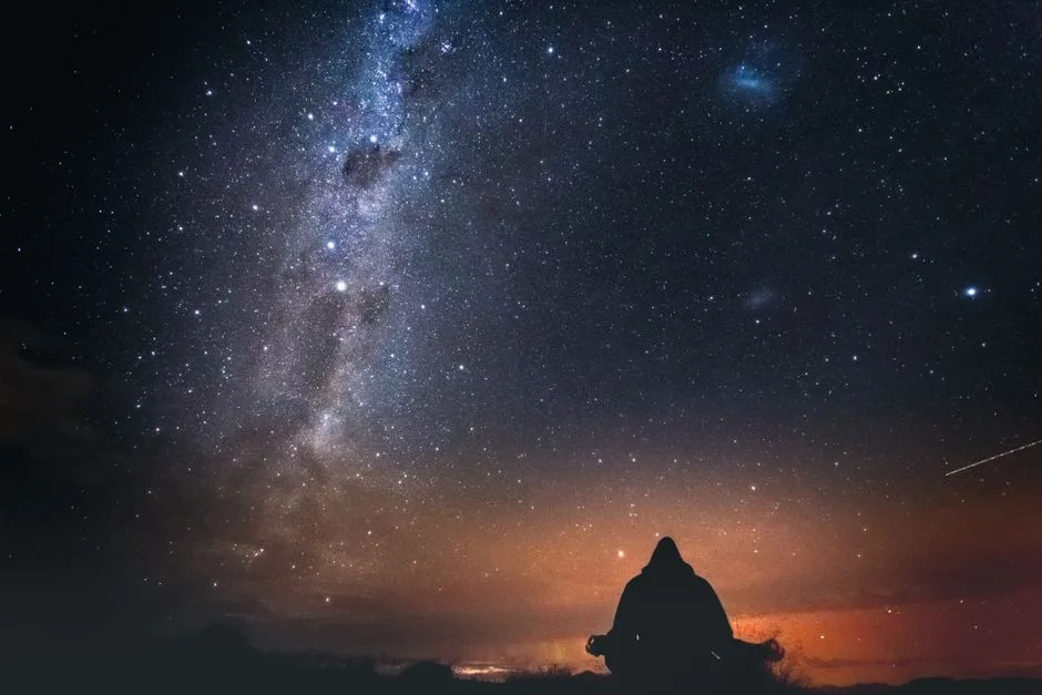 A lone figure sits under a starry sky with the Milky Way visible, capturing the beauty of a New Zealand night.