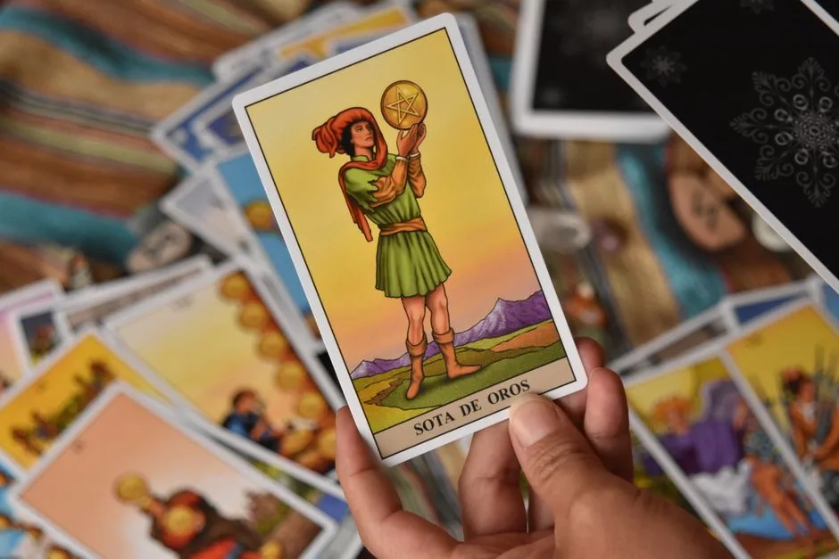 A person holds the Sota de Oros tarot card among a spread of cards for a reading.