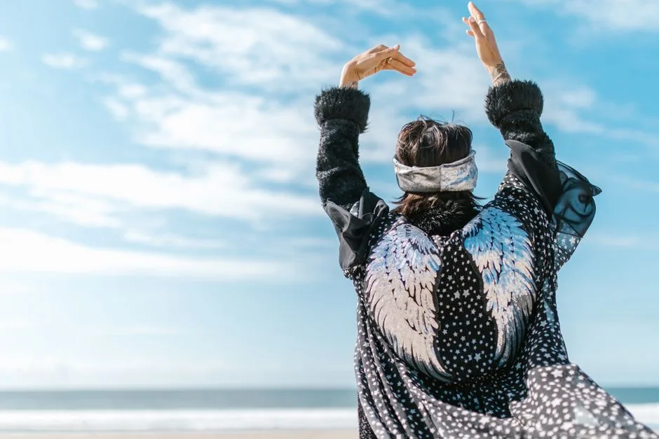 Person with angel wings raises arms in meditation on a sunny beach.