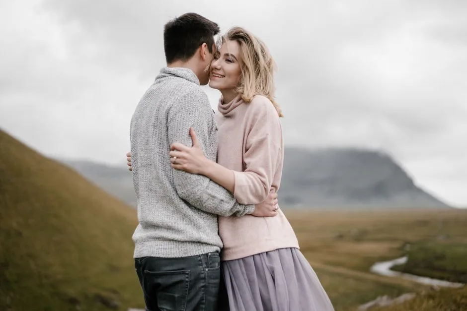 Side view cheerful embracing couple standing on autumn grassy hillside near narrow river and rocky mountains on foggy cold day