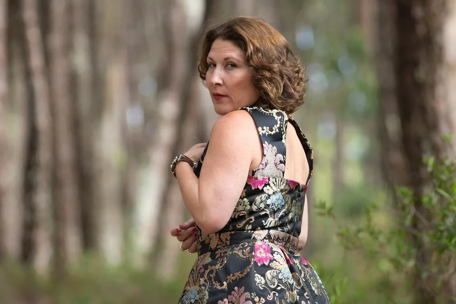 Woman in floral dress standing in a serene forest setting, looking over her shoulder.